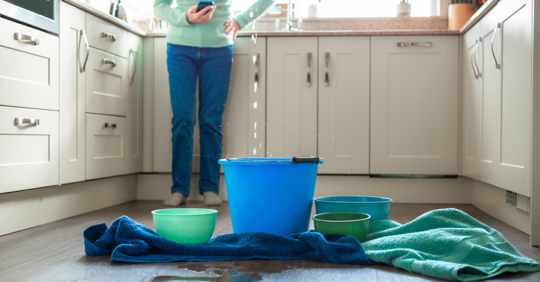 woman in kitchen with phone in her hand and a bucket catching water.